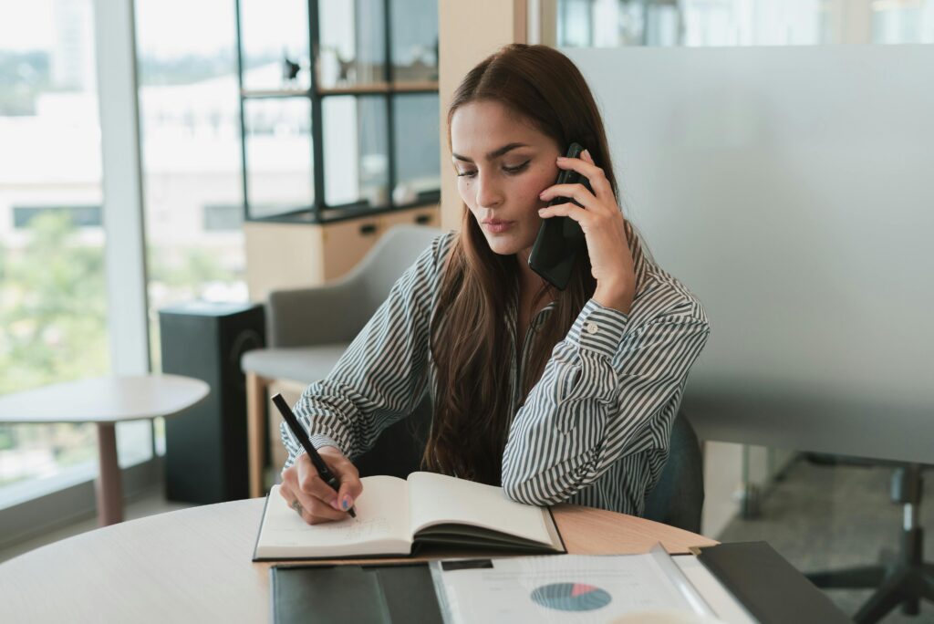 Young businesswoman on phone, jotting notes at stylish office desk.